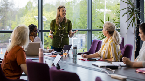 4 seated women listening to one woman who is standing in a meeting